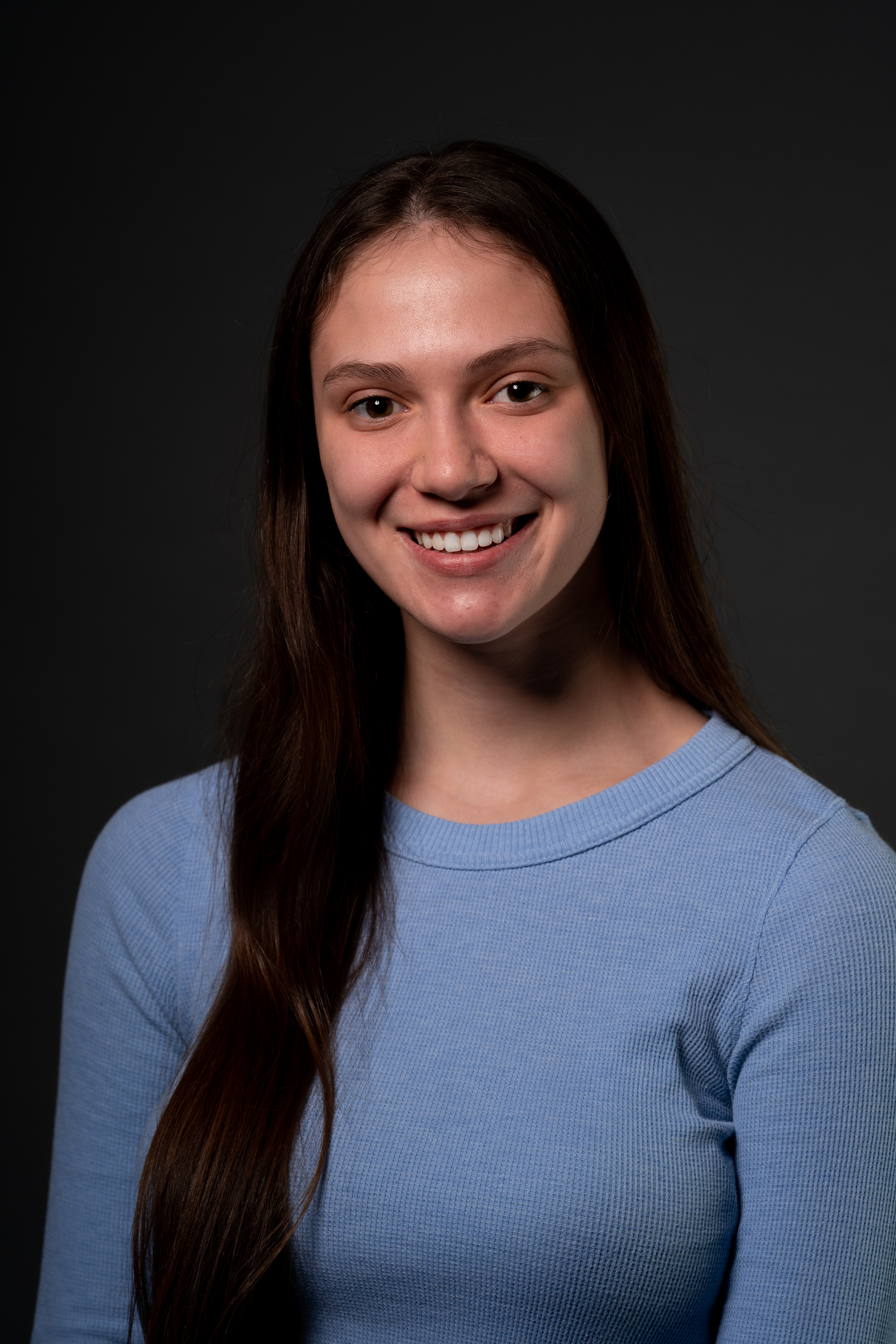 Headshot of Juliet Gentilucci. Juliet is wearing a blue long sleeved shirt and is smiling at the camera. She has olive-toned skin, long brown hair and hazel eyes. There is a dark gray background behind her.